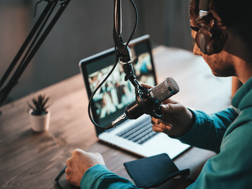 Wooden table with laptop on it - person sits at desk wearing headphones with overhead mic hanging down - recording a podcast