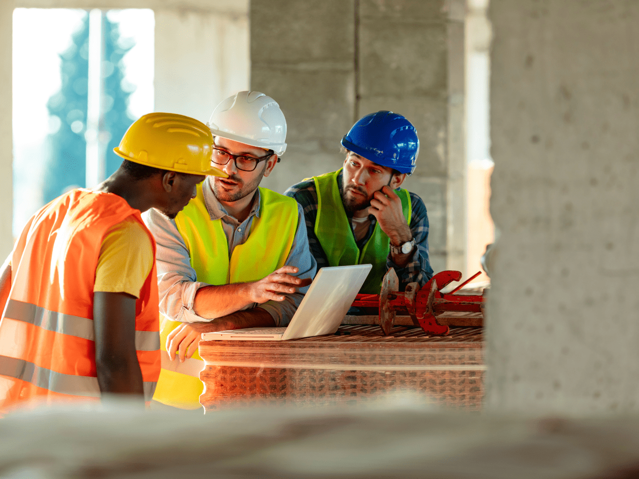 Three men in hi-vis uniforms, one wearing a blue hat, one wearing a white hat and the other wearing yellow all looking at a laptop at a construction site