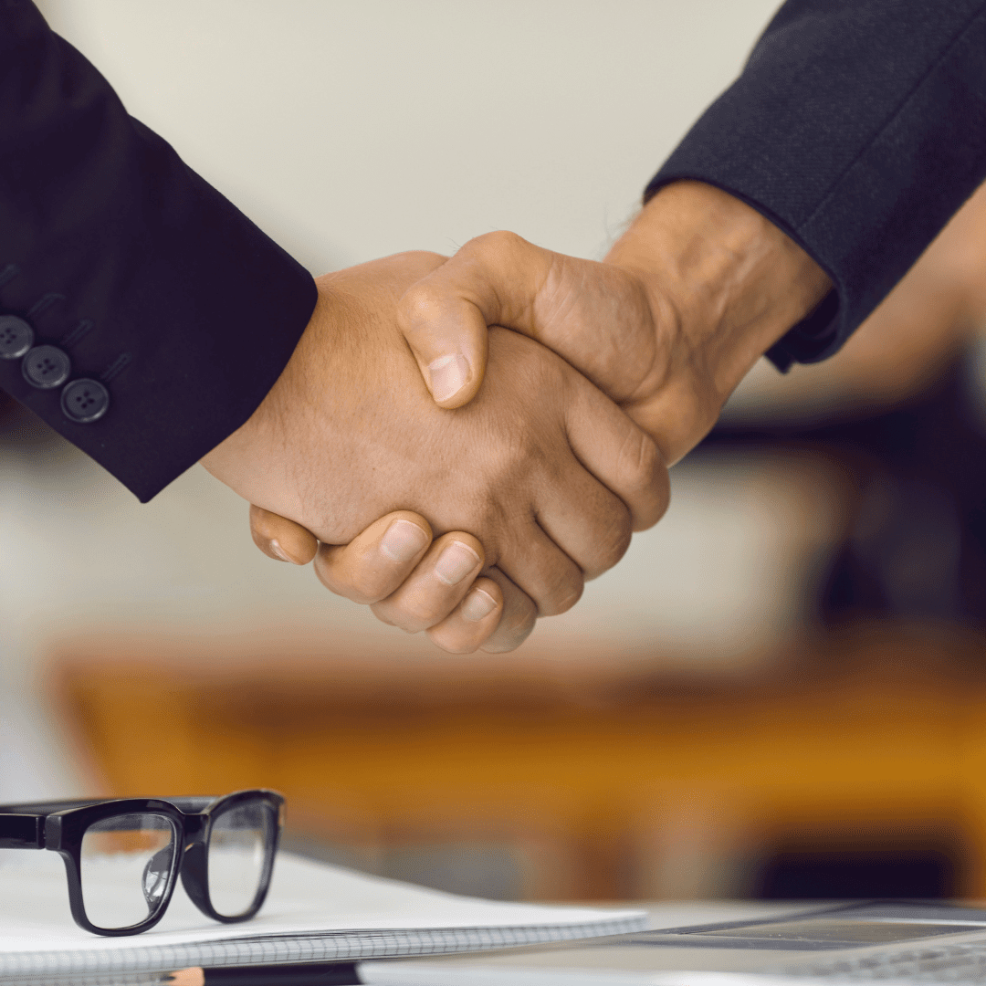 Two people shaking hands, there is paperwork under them shaking hands